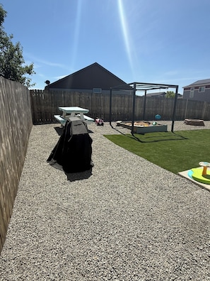 Ein Garten mit Holzzaun, Grill, Spielbereich mit Schaukel und einem kleinen Bereich mit Rasenteppich.
