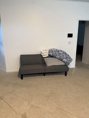 Ein graues Sofa mit einer gefalteten Decke und Kissen vor einer weißen Wand, eine Steckdose und eine Tür, die in einen anderen Raum führt.
