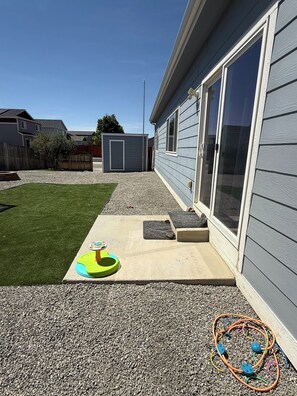 Ein Wohnhausgarten mit Rasenfläche, einer kleinen Spielzone inklusive Spielzeug und einem Kiesweg, der zu einem Haus mit Glastür führt.