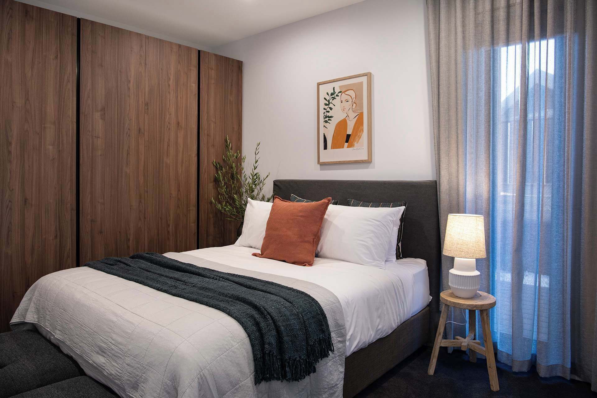 A hotel room with a bed, a nightstand, a lamp, a window with curtains, and a wall-mounted artwork.