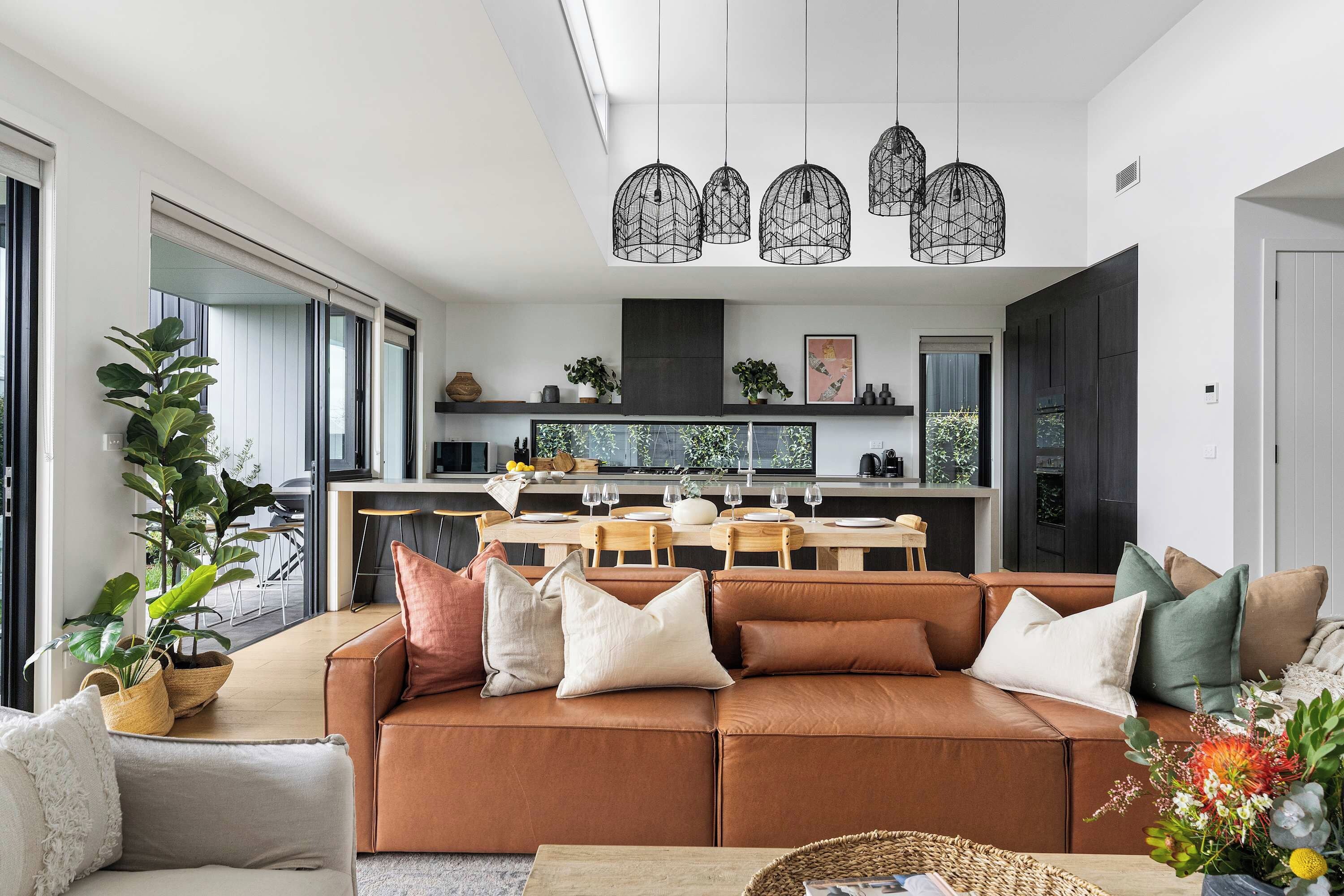 A modern living room with a leather sofa, a dining area with a kitchen in the background, and a large window providing natural light.