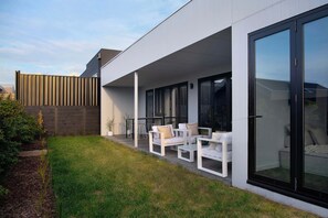 Un patio moderno al aire libre con mobiliario blanco y un ventanal de vidrio que da a un área cubierta.