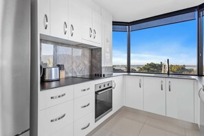 A modern kitchen with white cabinets, a black countertop, and a view of the outdoors through a large window. - Esplanade Views with Pool, CBD (Cairns North)