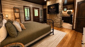 A bedroom with a bed, a wooden ceiling fan, a stone accent wall, a mounted TV, and a visible bathroom with a sink and mirror. - Luxury cabin w/ Hot Tub, Pool Table, Arcade & Stone Soaking Tub - Broken Bow, OK (Broken Bow)
