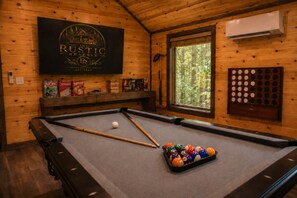 A pool table with a cue stick, pool balls, and a window with a view of trees. - The Rustic Reserve – Luxury cabin with Hot Tub, Pool Table, Arcade & Stone Soaking Tub - Broken Bow, OK (Broken Bow)