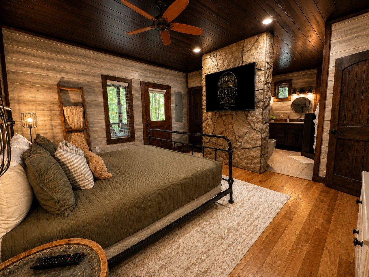 A bedroom with a bed, a wooden ceiling fan, a stone accent wall, a television, and a view of a bathroom.