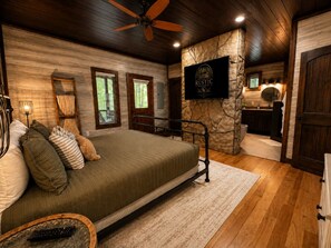 A bedroom with a bed, a wooden ceiling fan, a stone accent wall, a television, and a view of a bathroom. - The Rustic Reserve – Luxury cabin with Hot Tub, Pool Table, Arcade & Stone Soaking Tub - Broken Bow, OK (Broken Bow)