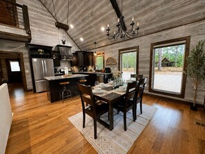 A spacious kitchen with wooden floors, a dining area with a table set for four, and large windows offering a view of the outdoors. - The Rustic Reserve – Luxury cabin with Hot Tub, Pool Table, Arcade & Stone Soaking Tub - Broken Bow, OK (Broken Bow)