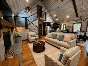 A spacious living area with a fireplace, a sofa set, a coffee table, and a staircase leading to an upper level. - The Rustic Reserve – Luxury cabin with Hot Tub, Pool Table, Arcade & Stone Soaking Tub - Broken Bow, OK (Broken Bow)
