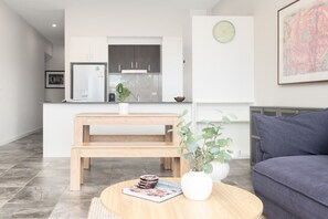 A modern kitchen with white cabinets, a refrigerator, and a sink. There is a wooden bench and a round table with a plant and a magazine. A gray sofa is in the foreground. - Quiet Turner Stay | Walk to Braddon & ANU (Turner)