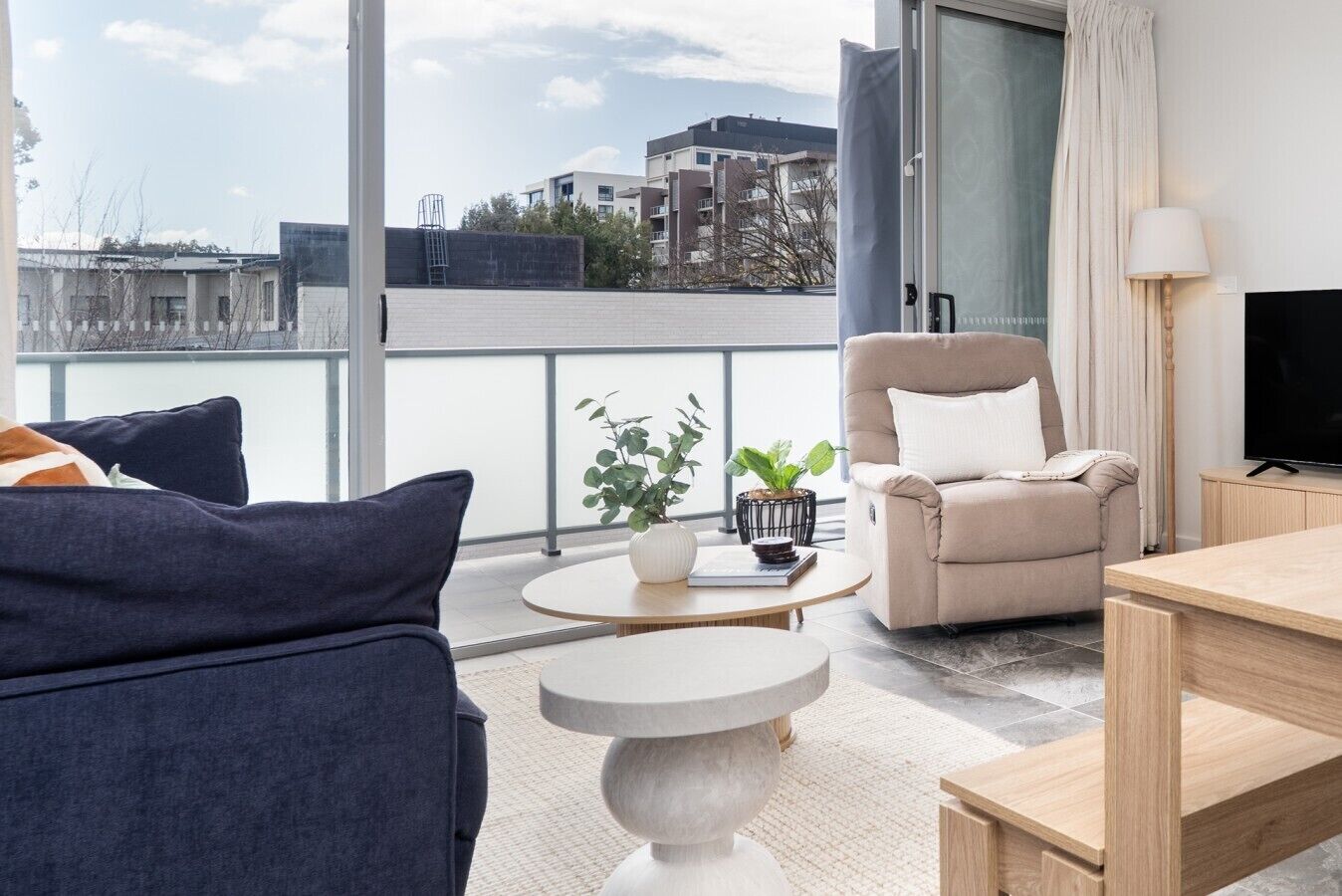 Un salon moderne comprenant un canapé, un fauteuil et une table basse, avec une vue sur la ville grâce à de grandes fenêtres.
