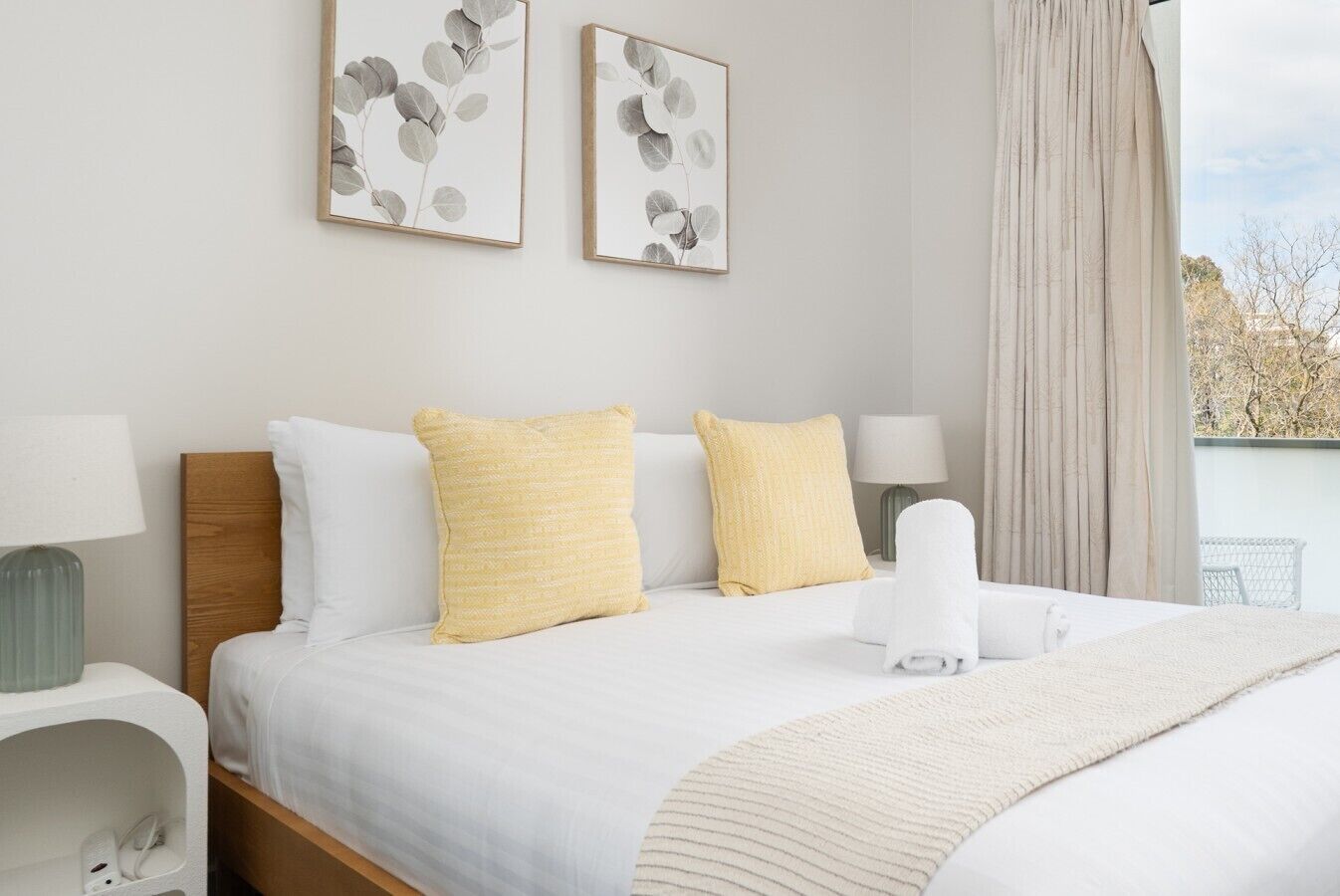 A hotel room with a bed, two pillows, a blanket, a bedside table with a lamp, and a window with curtains.