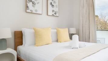 A hotel room with a bed, two pillows, a blanket, a bedside table with a lamp, and a window with curtains.