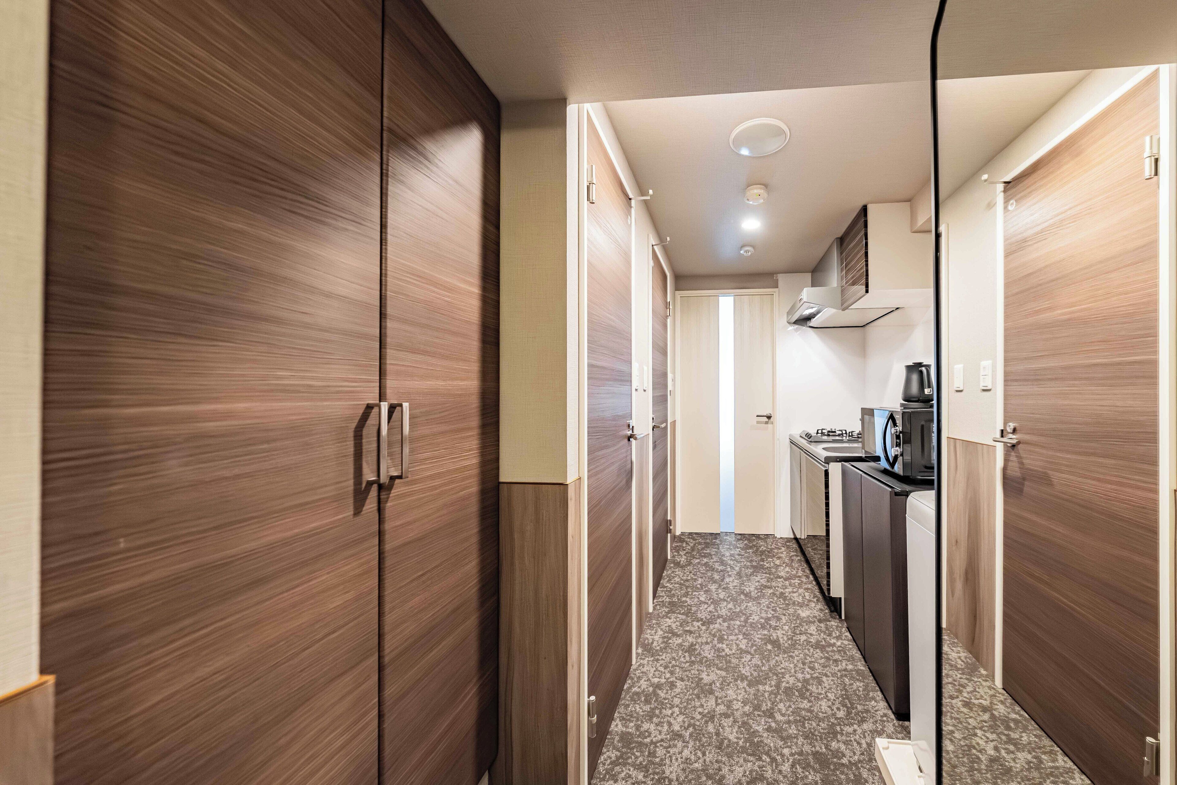 A hallway with wooden wardrobes, a kitchen area with a stove, and a microwave.