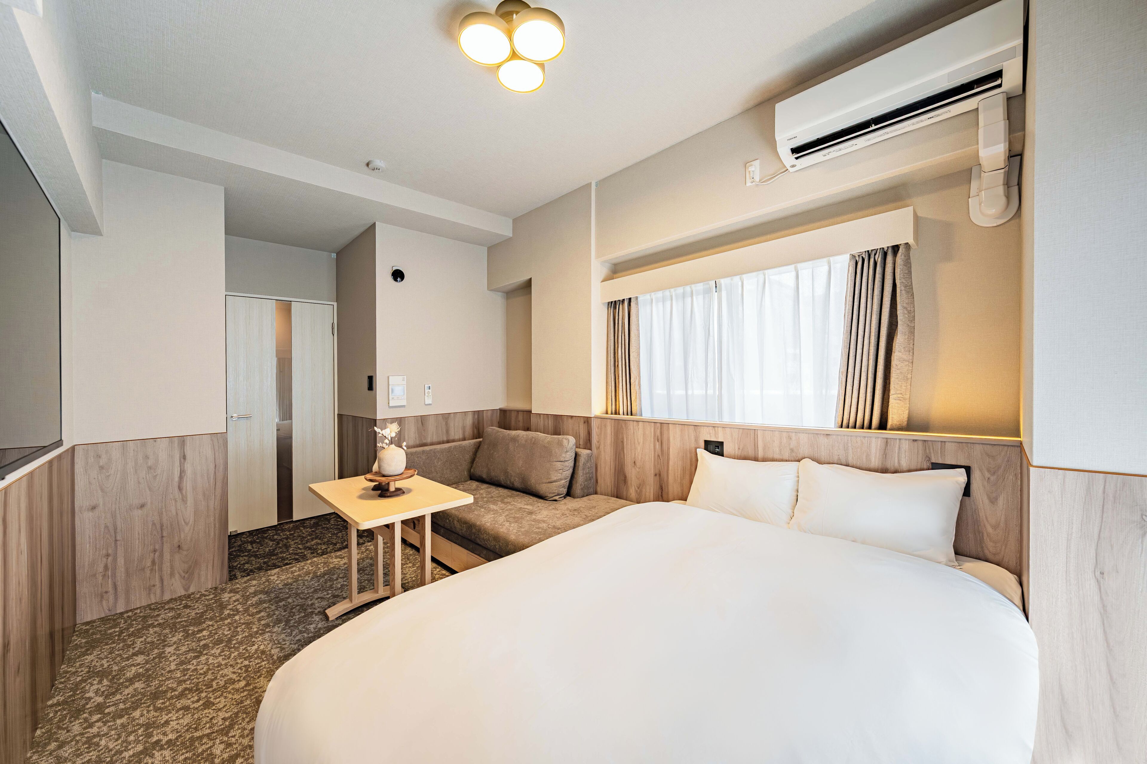 A hotel room with a large bed, a small table, a sofa, and a window with curtains.