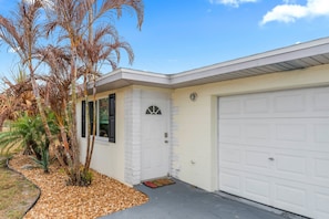 Ein eingeschossiges Haus mit Garage, einer kleinen Veranda und einem Garten mit Palmen und einem Kiesweg.