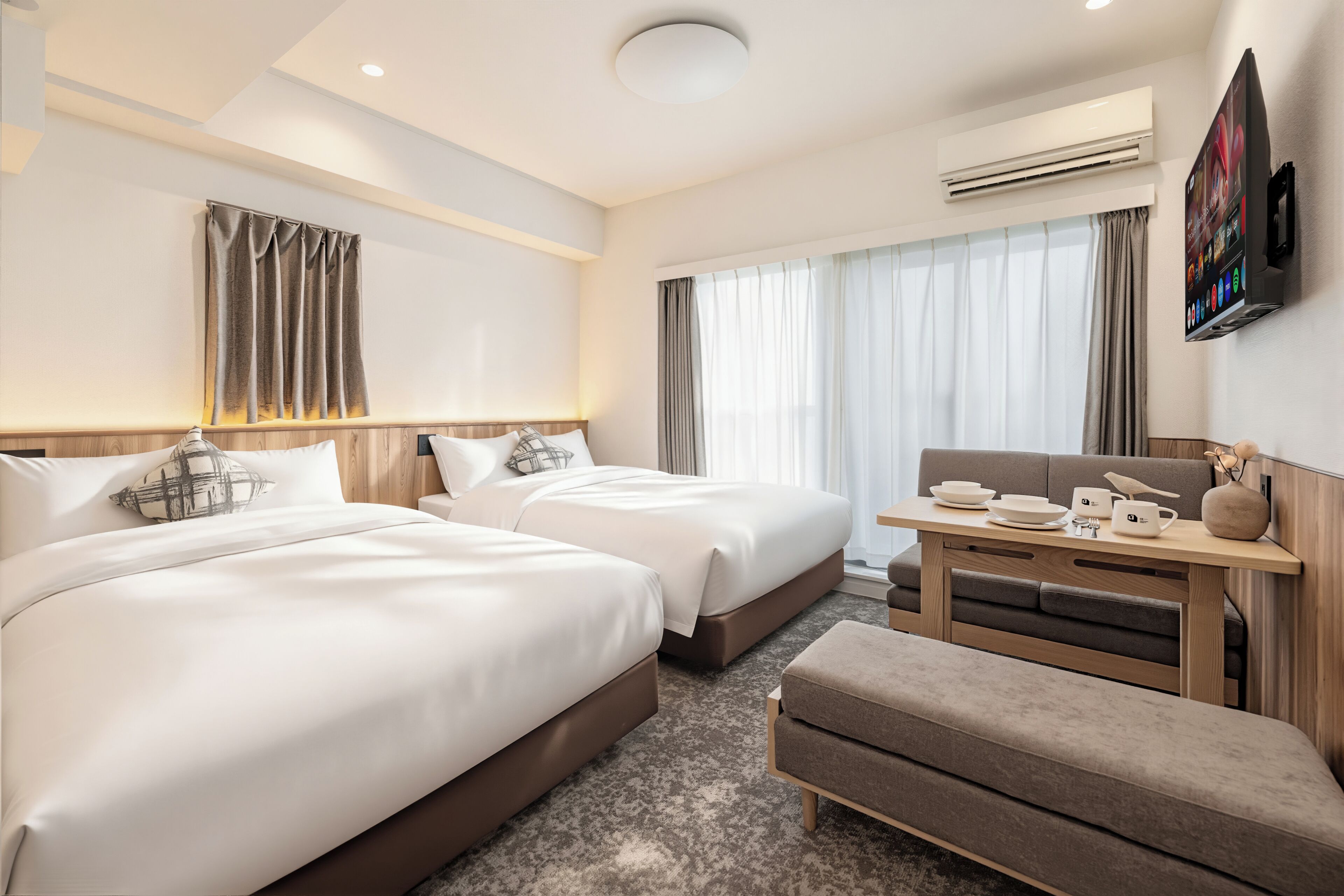 Une chambre d’hôtel avec deux lits, une petite table avec une chaise, un téléviseur à écran plat fixé au mur et un appareil de climatisation.