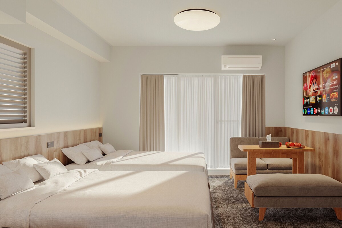 A hotel room with two beds, a desk, a chair, a small table, and a wall-mounted TV.