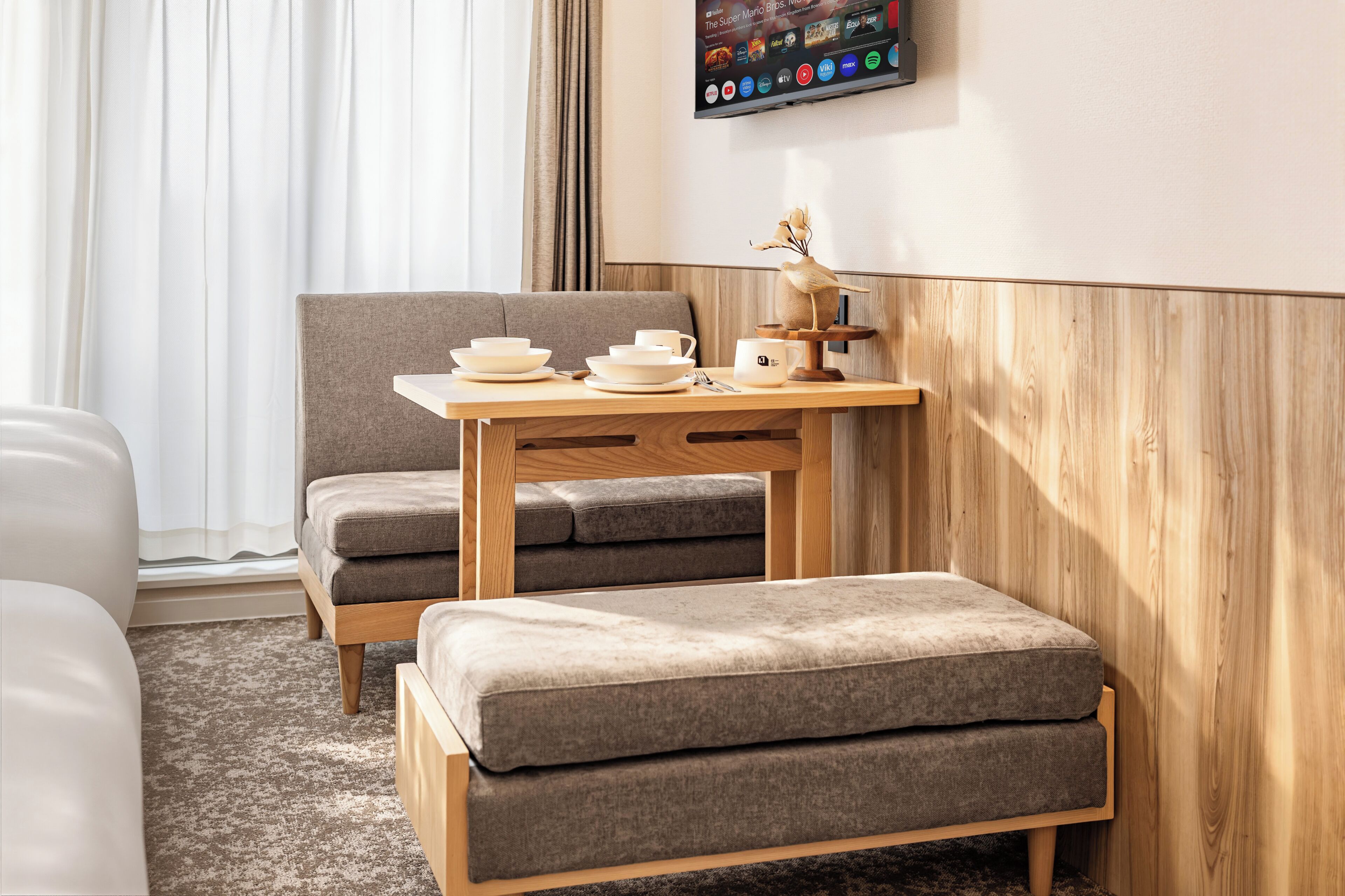Une chambre d’hôtel moderne avec une table en bois, deux fauteuils gris rembourrés, un petit tabouret en bois et un téléviseur fixé au mur qui affiche différents services de diffusion en continu.