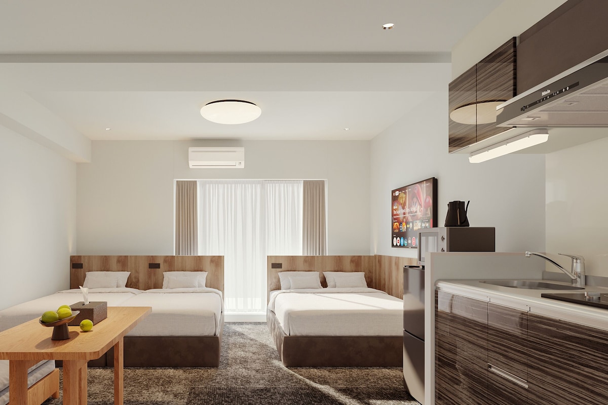 A compact hotel room with a bed, a small table with fruits, a sofa, a kitchenette, and a large window with curtains.