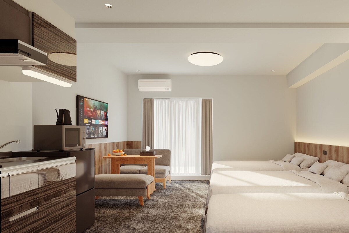 A modern hotel room with a kitchenette, a flat-screen TV, a dining area, and a bed with a wooden headboard.