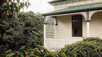 A white building with a green roof, a white porch with a lattice railing, and dense green foliage in the foreground.