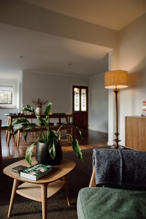 A living room with a sofa, a coffee table with a book, a potted plant, a lamp, and a dining area in the background.
