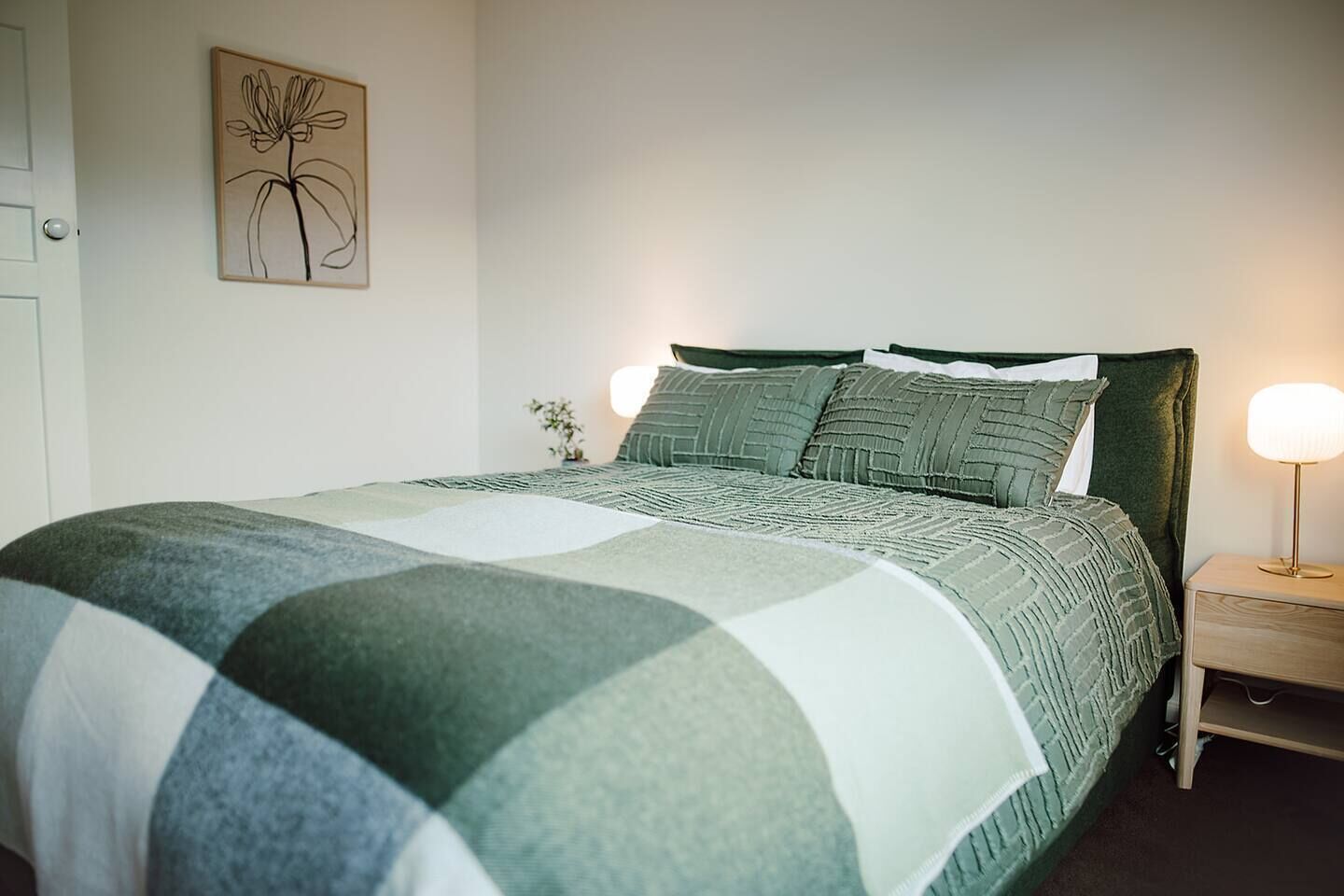 A neatly made bed with a green and white checkered bedspread, a wooden nightstand with a lamp, and a wall-mounted picture of a flower.