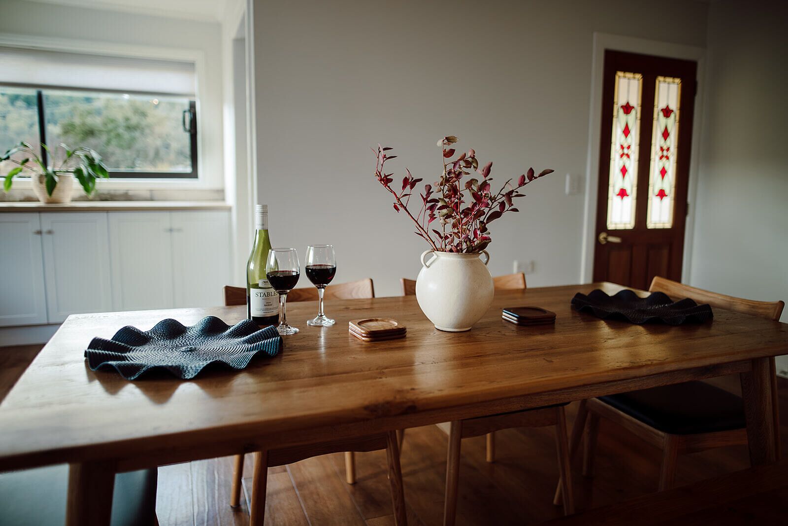 Una mesa de comedor con un florero de plantas de hojas rojas, dos copas de vino, una botella de vino y un mantel individual tejido.