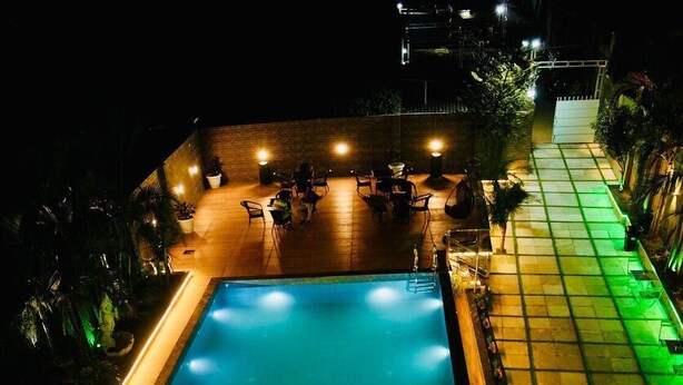 A rectangular swimming pool with a wooden deck surrounding it, illuminated by overhead lights. - MH Club And Resorts (Nagri)