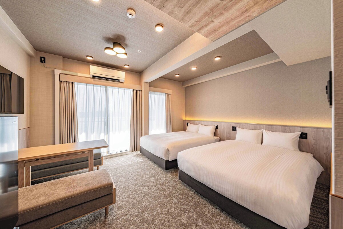 A hotel room with two beds, a desk, a bench, and an air conditioning unit.