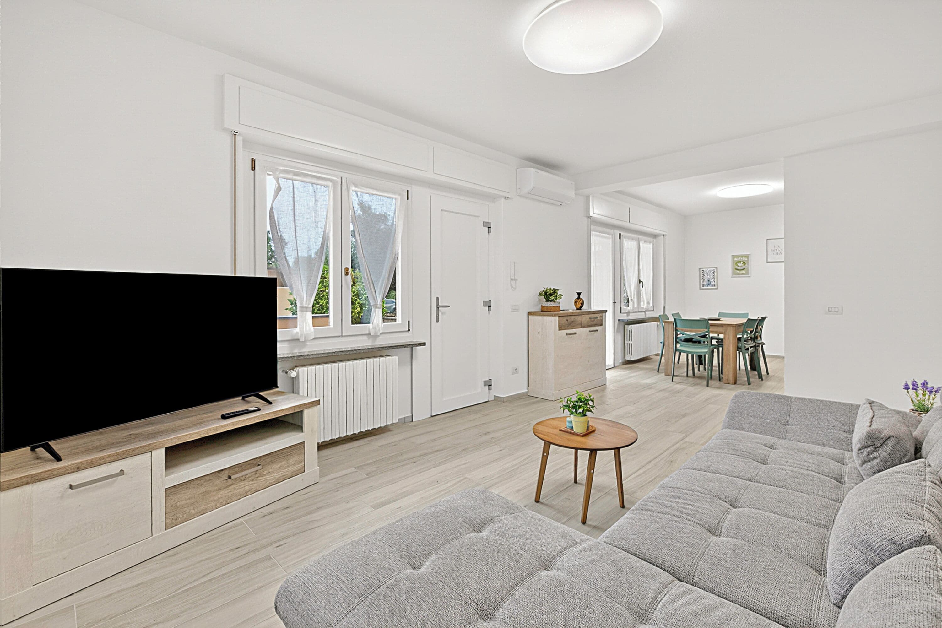 A modern living room with a flat-screen TV, a grey sofa, a small wooden coffee table, and a dining area with a table and chairs.