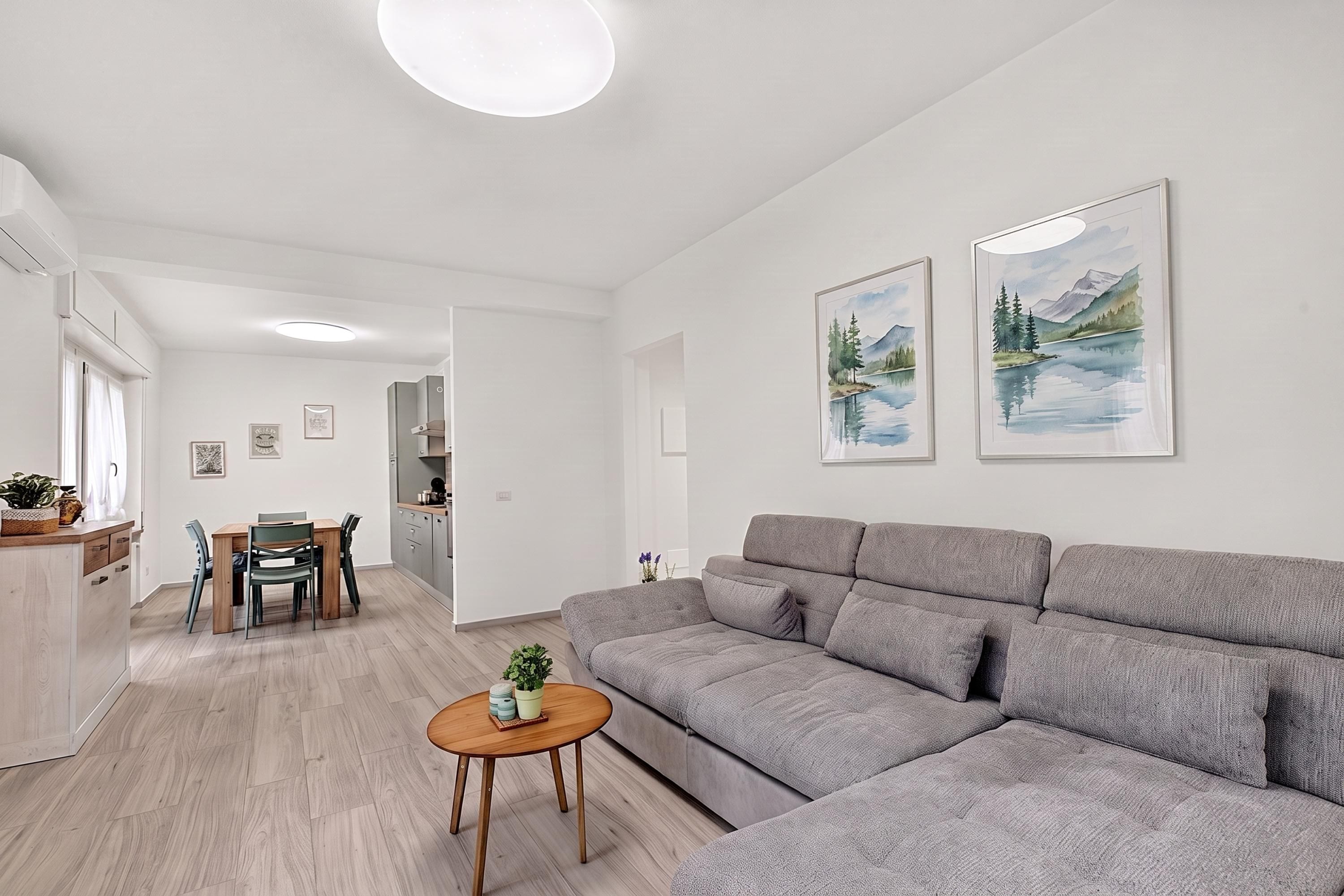 Uma sala de estar moderna com um sofá cinza, uma mesa de centro de madeira e uma área de jantar com mesa e cadeiras. Há duas pinturas de paisagens emolduradas na parede.