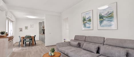 Uma sala de estar moderna com um sofá cinza, uma mesa de centro de madeira e uma área de jantar com mesa e cadeiras. Há duas pinturas de paisagens emolduradas na parede.
