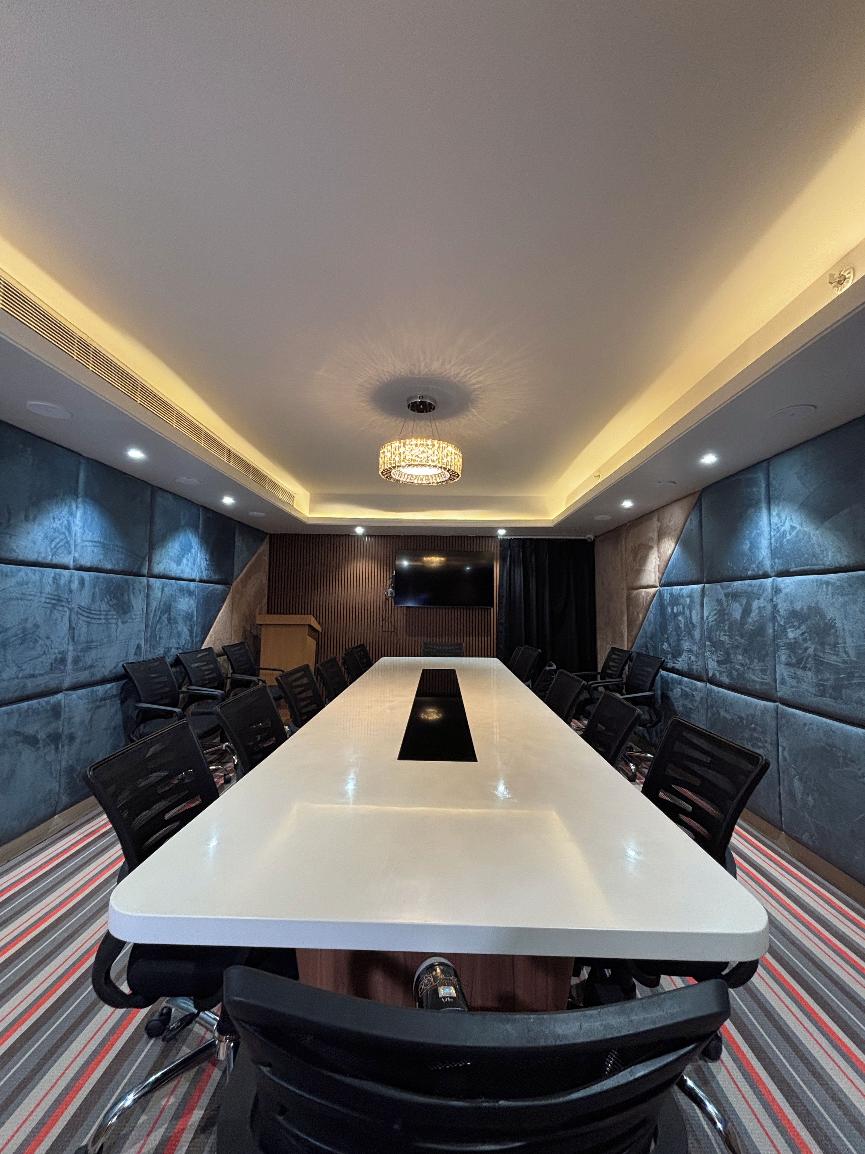 A conference room with a long table, chairs, a television screen, and a patterned carpet.