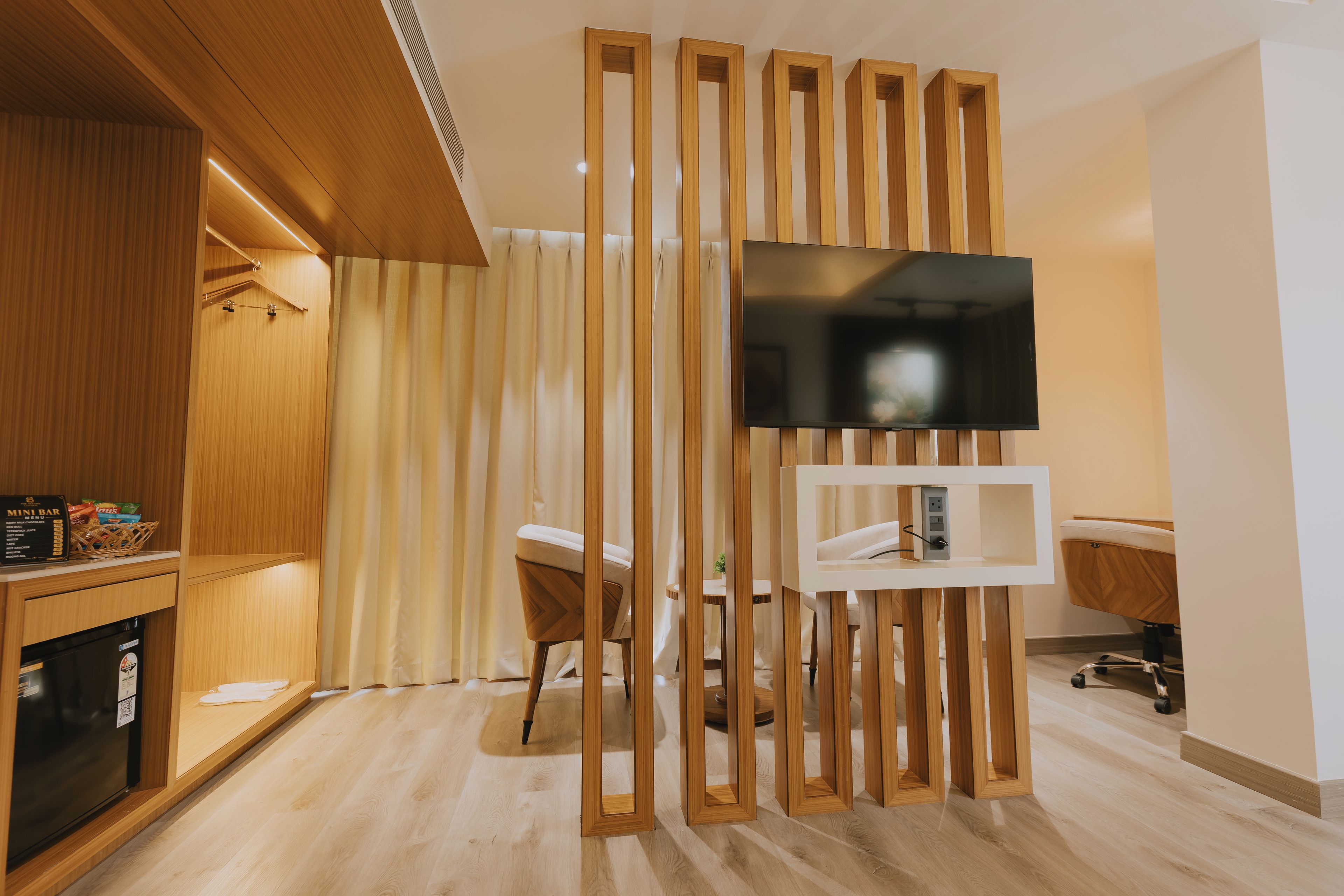 Habitación de hotel moderna con un separador de madera, un televisor de pantalla plana sobre un soporte, un minibar y una zona de estar con una silla y una pequeña mesa.