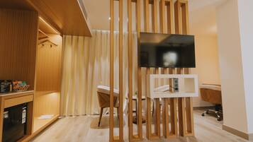 Habitación de hotel moderna con un separador de madera, un televisor de pantalla plana sobre un soporte, un minibar y una zona de estar con una silla y una pequeña mesa.
