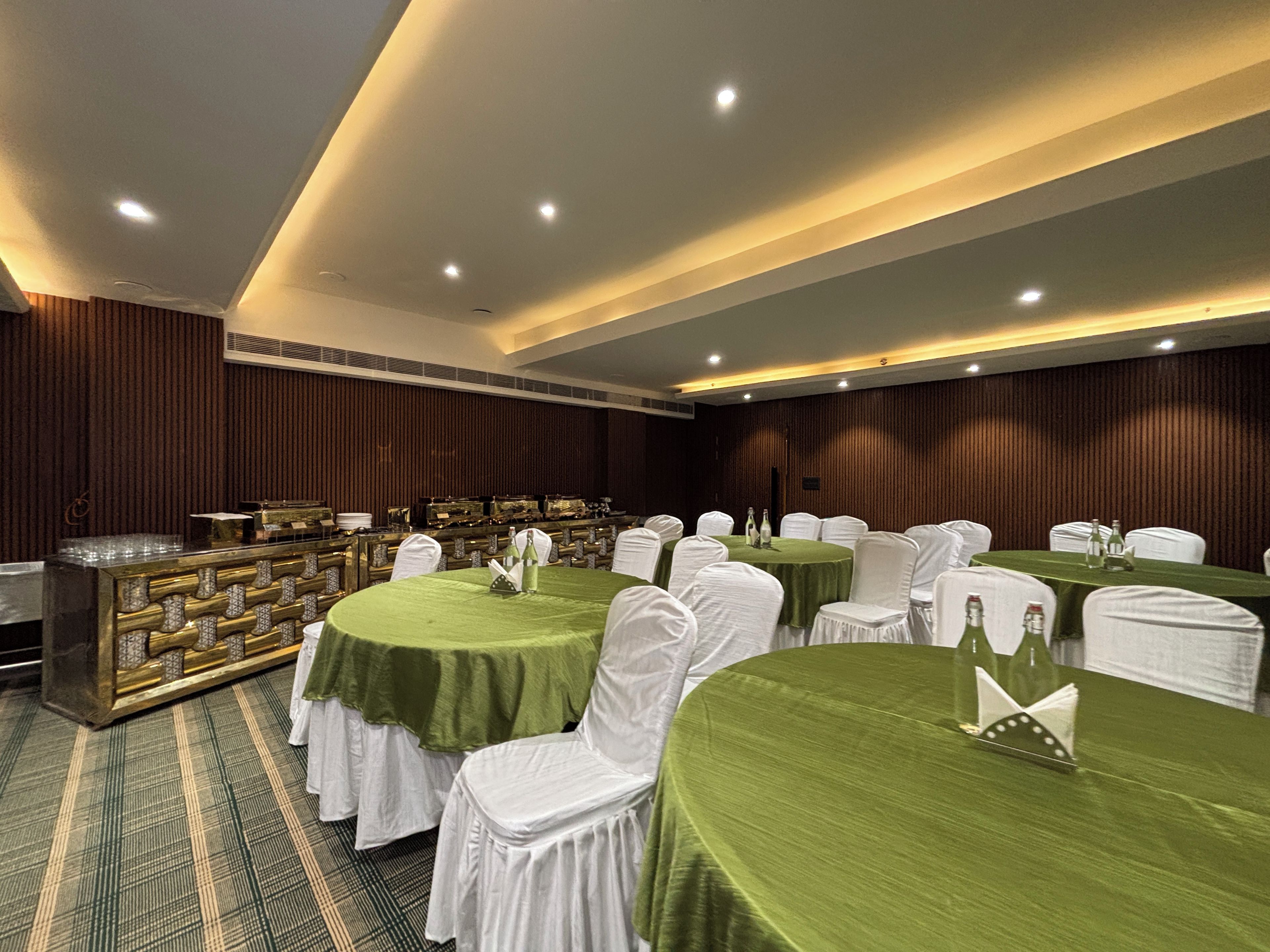 Un salón de banquetes con mesas redondas cubiertas con manteles verdes, sillas blancas y una zona de buffet con un mostrador dorado.