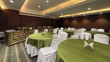 Un salón de banquetes con mesas redondas cubiertas con manteles verdes, sillas blancas y una zona de buffet con un mostrador dorado.