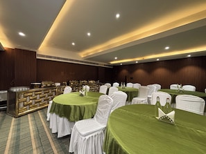Un salón de banquetes con mesas redondas cubiertas con manteles verdes, sillas blancas y una zona de buffet con un mostrador dorado.