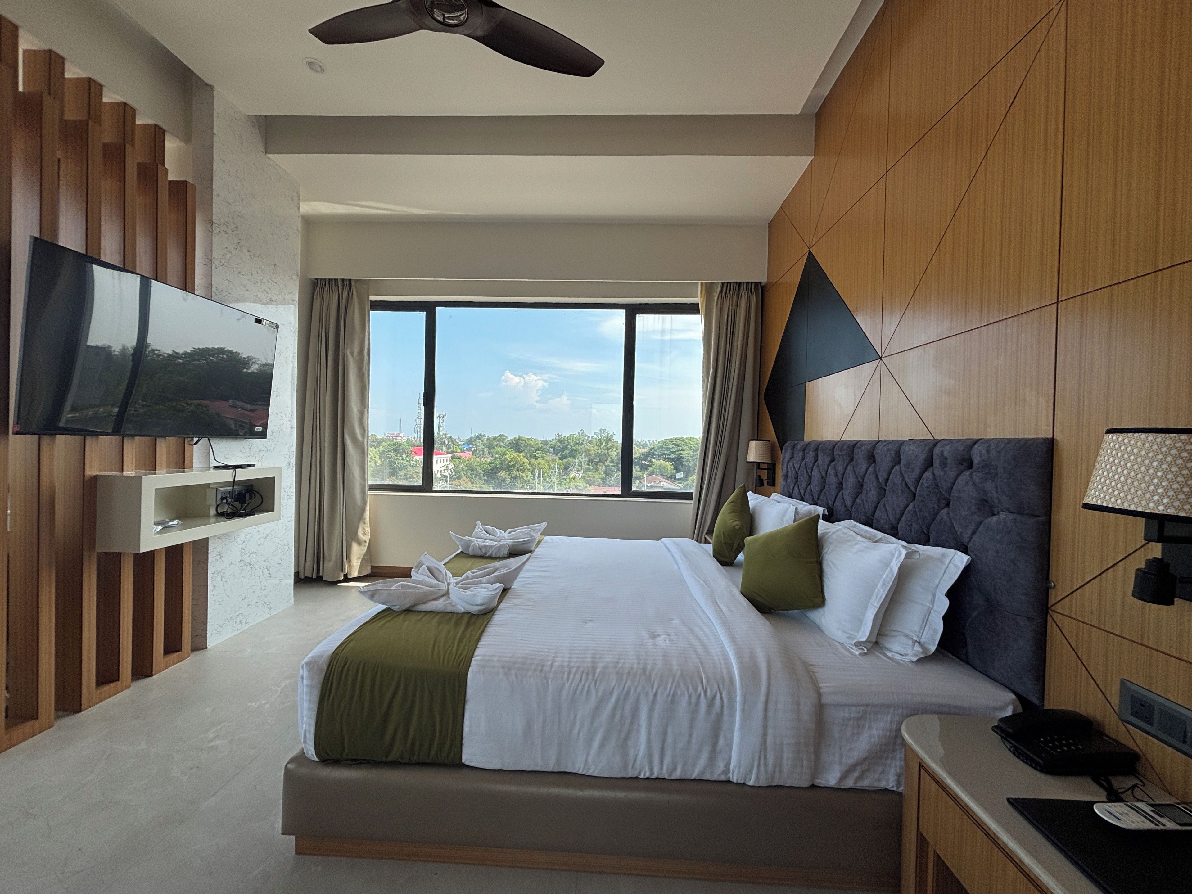 Una habitación de hotel moderna con una cama grande, un televisor de pantalla plana montado en la pared, un escritorio con lámpara y vistas a la ciudad a través de la ventana.