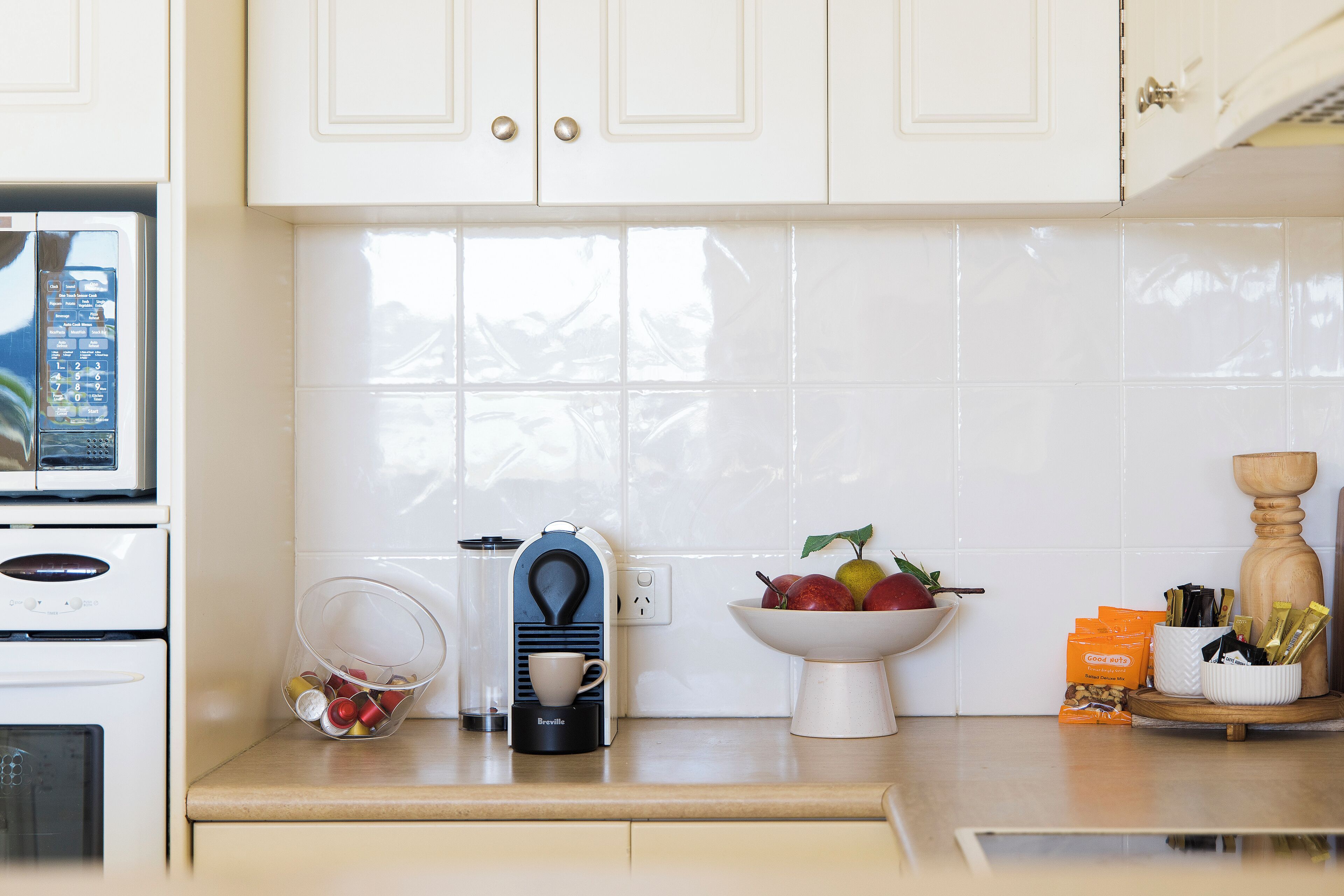 A kitchen with white cabinets, a Breville coffee maker, a bowl of fruit, and a coffee machine.