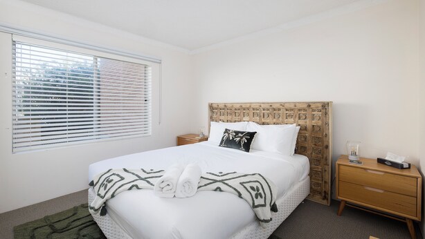 A hotel room with a bed, a nightstand, a bedside table, a window with blinds, and a headboard with a decorative pattern. - Bella Vista Shoal Bay (Shoal Bay)