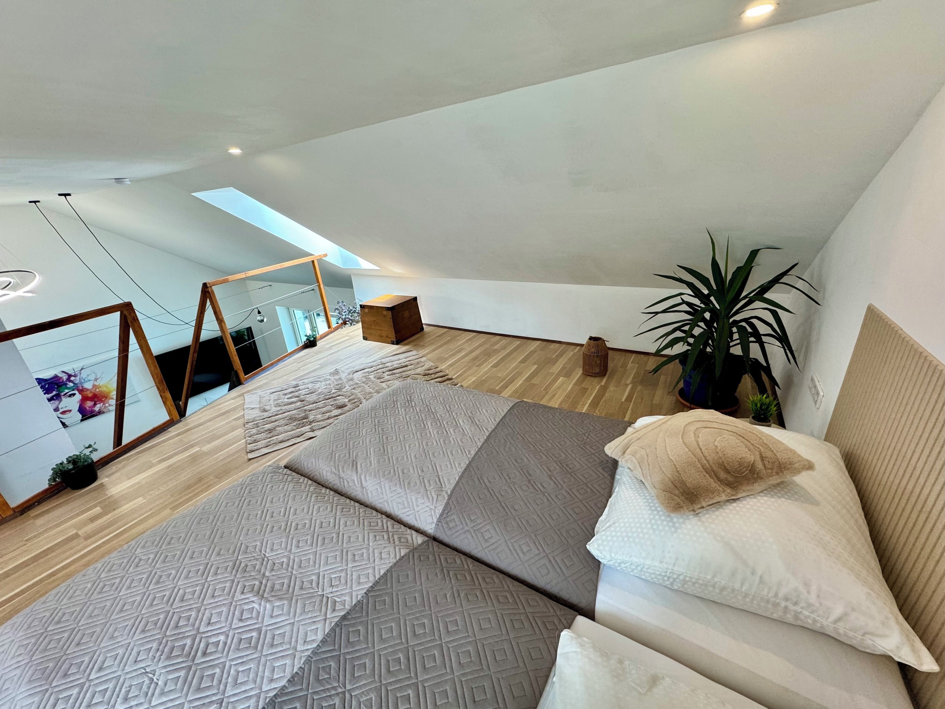 Un dormitorio moderno con techo inclinado, una cama grande con ropa de cama estampada, una planta en maceta y vistas de la ciudad a través de un tragaluz.