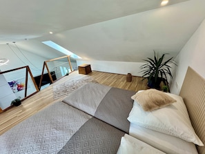 A modern bedroom with a sloped ceiling, a large bed with patterned bedding, a potted plant, and a view of the city through a skylight.