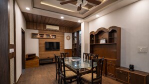 A dining area with a glass table surrounded by chairs, wooden cabinets, a television mounted on the wall, and a ceiling fan.
