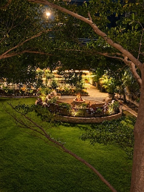 A well-lit garden area with a central fire feature, surrounded by various plants and a seating area.