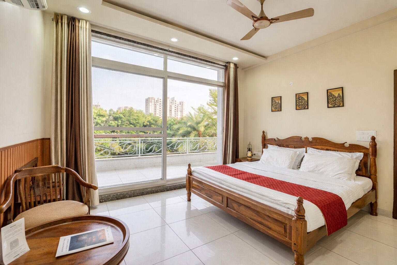 A hotel room with a large bed, a wooden chair, a small table, and a balcony with a view of the city.