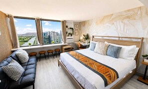 A modern bedroom with a large bed, a sofa, a desk, and a view of a cityscape. - Waikiki 2BR 2BA 34th Fl Ocean View Sunset/Sunrise (Honolulu)