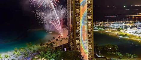 Korkea hotellirakennus, jossa on uima-allas ja näköala rannalle, ja jonka yllä ilotulitus valaisee taivaan.